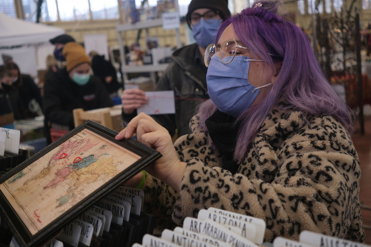 A woman with purple hair is dressed in an animal print coat. She is smiling under her mask as she shows an embroidered map to a customer at the Christmas market.