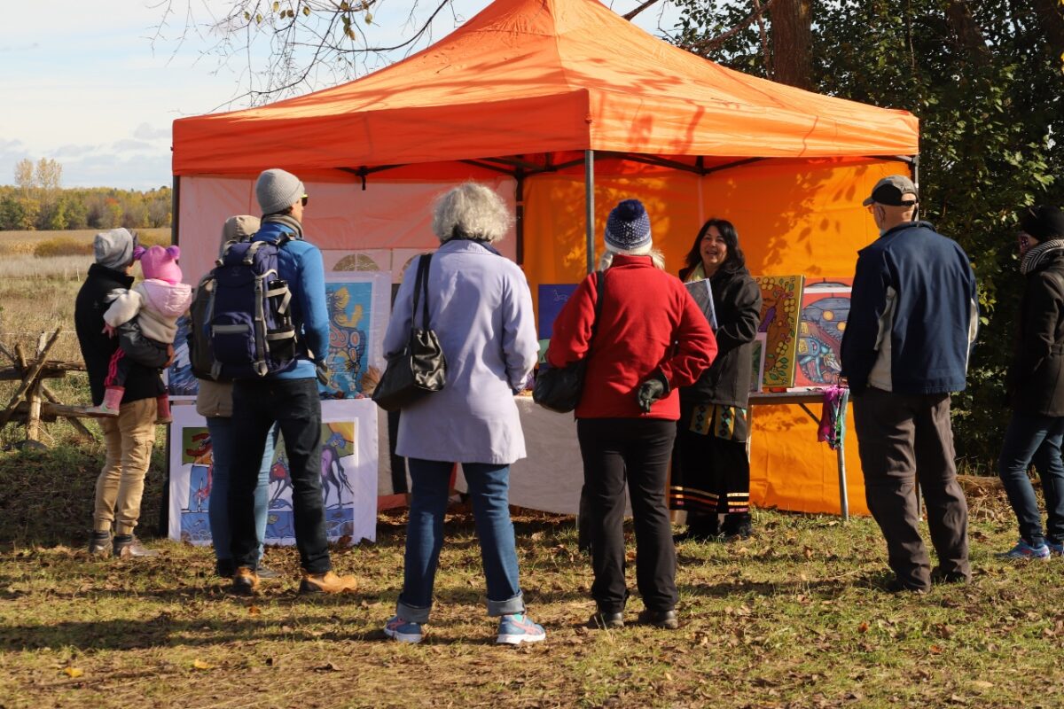 Spirit horse caretaker and artist Rhonda Snow is talking with festival attendees about her spirit horse herd and showing her paintings to visitors at her vibrant orange tent. 