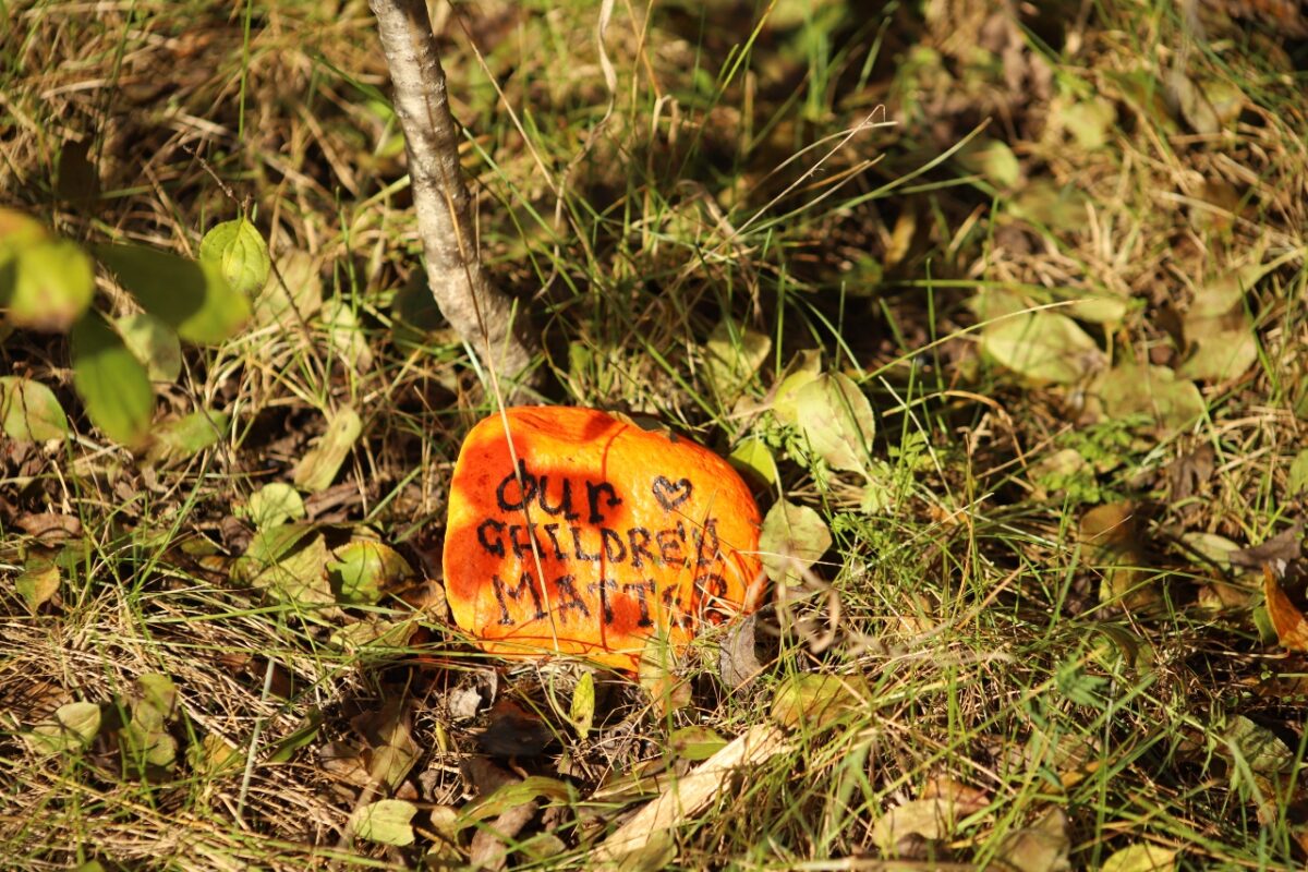 One of the orange reconciliation rocks is pictured in the grass near a small tree with the words 'our children matter' in black. 