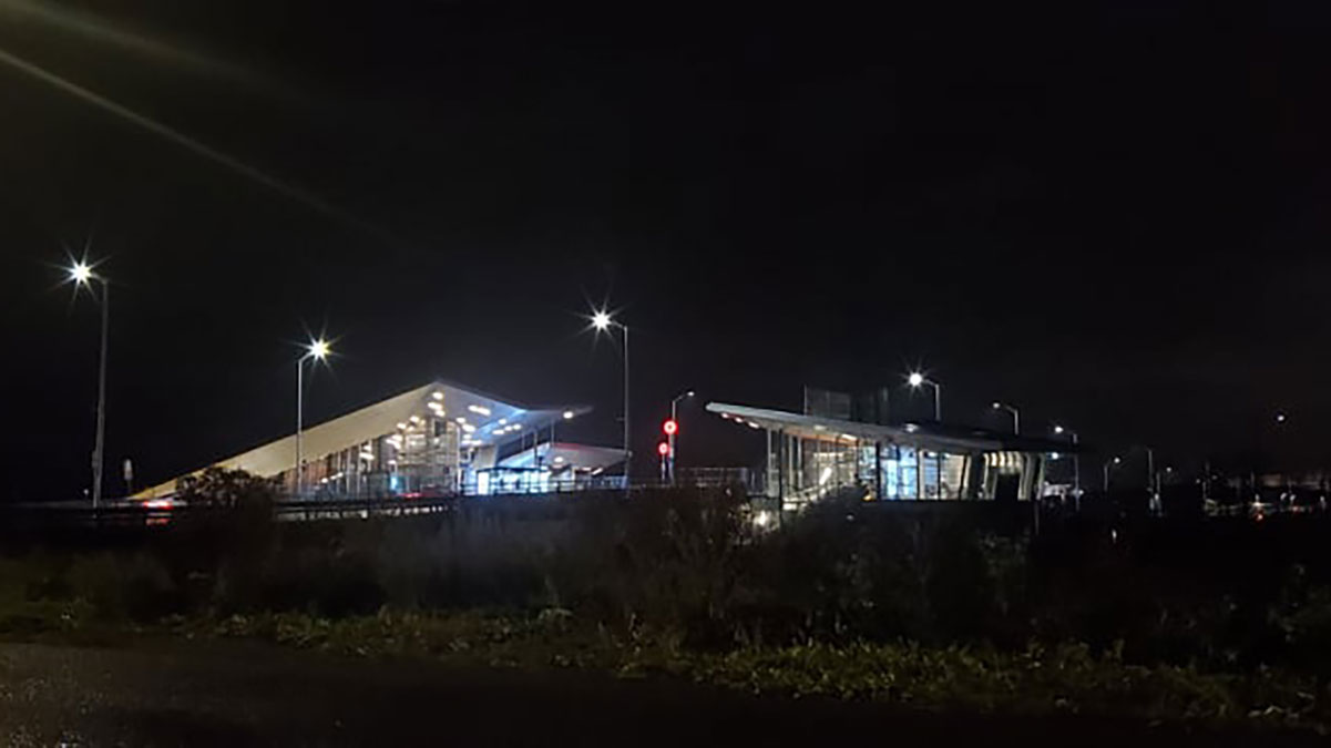 The Pimisi train station is shown at night, with grass in the foreground and a car drives near the station's two platforms