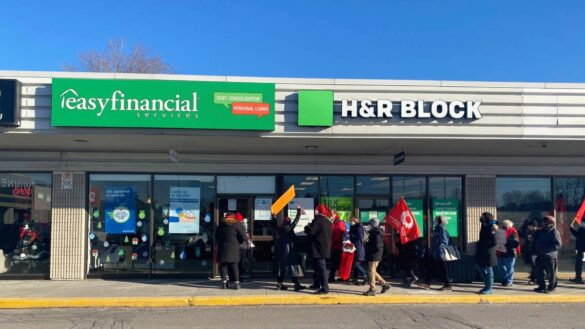 A group of advocates march with red flags towards a loan agency with a green sign.