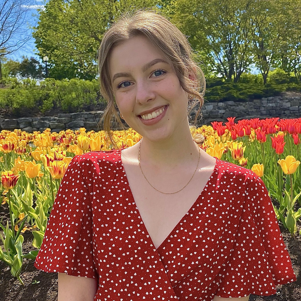 A young white woman with blonde hair sits in a field of yellow and red tulips with a red dress on. She's smiling at the camera.