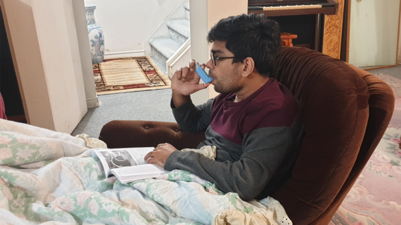 A man, sitting on a chair and wrapped in a blanket, is using a metered dose inhaler.