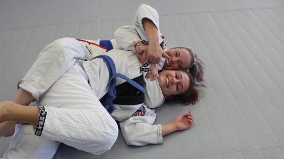 Two girls training Brazilian Jiu Jitsu together, wearing white gis with blue belts on a grey mat.