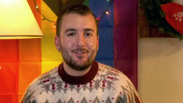 Reporter Ben Sylvestre sits in front of a Progressive Pride Flag. He wears a holiday sweater.