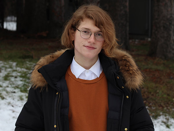Nat Jackett, wearing an orange sweater, white button-down and black jacket, stands in front of a forested area on a half-snowy field