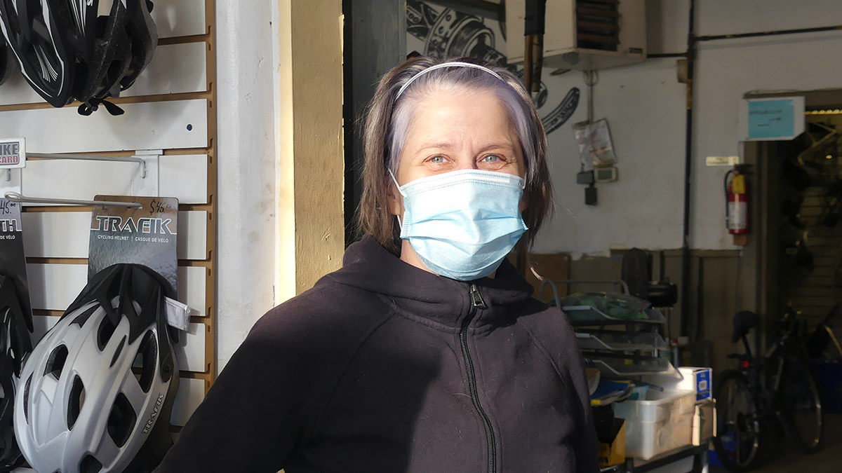 Torunn Gill, in a black hoodie, blue mask and white headband, stands next to a wall with bike helmets for sale
