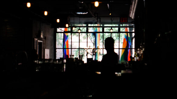 Silhouette of someone in a dark room, standing in front of window panes through which pride flags can be seen
