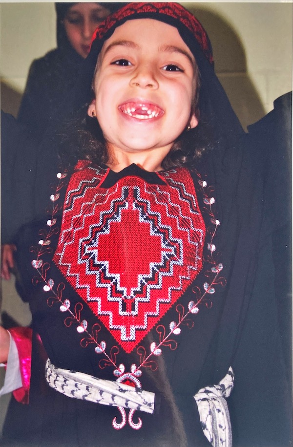 Young girl smiling widely while wearing black dress with large, red tatreez pattern