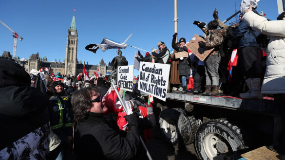 Protesters gather near parliament as part of the “Freedom Convoy” that occupied Ottawa in January. Protesters are gathered on a truck bed and on the ground. Many are wearing winter coats and holding signs. Parliament is seen in the background.