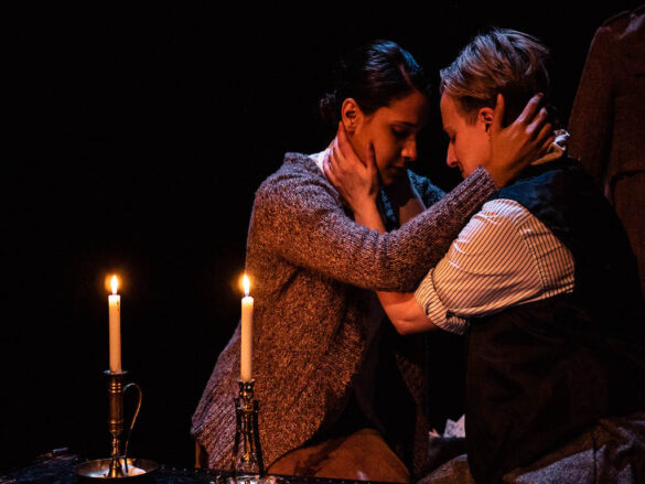 Two actors embrace one another on a dimly lit onstage. There are candles set on the left side of the stage.