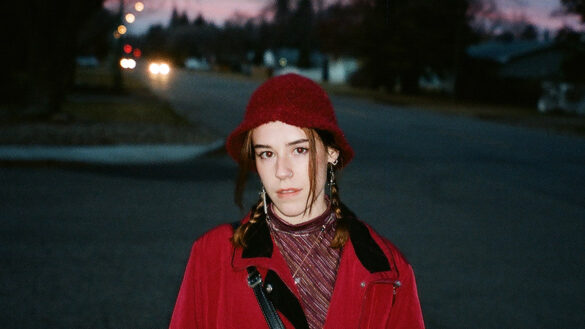 A young brunette woman in a red bucket hat and jacket stands in front of a sunset on a suburban street.