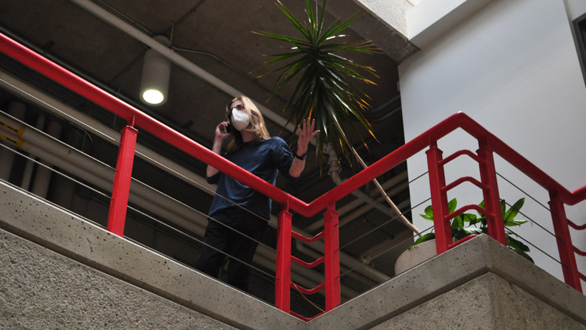 A blonde man talks on the phone with his hands extended. He is standing on a balcony with a red hand-rail.
