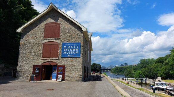 A stone building with a blue sign that says Bytown Museum has open wooden doors. The Rideau Canal is on the right with boats waiting on the locks.