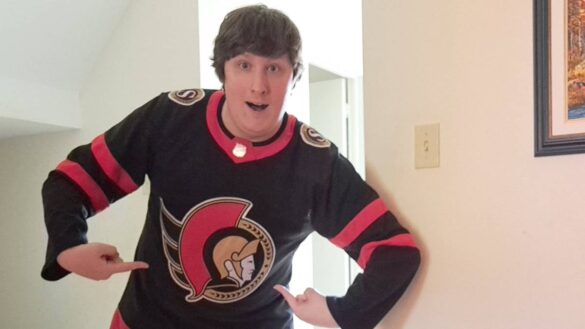 Matt Bostelaar wears a black Ottawa Senators jersey. He points to the crest with both hands.