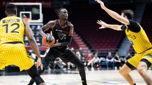 Deng Adel looks towards the basket holding the basketball in a black Ottawa BlackJacks jersey. Two Hamilton defenders wear yellow jerseys and are guarding Adel.