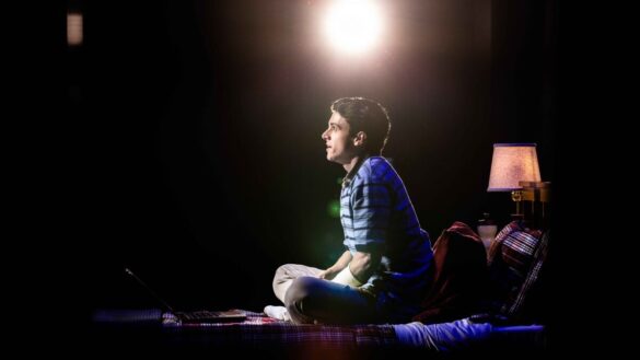 The character Evan Hansen sits with his legs crossed and looks ahead.