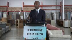 Minister Ahmed Hussen stands at a podium saying "Reaching Home" in a building with furniture.