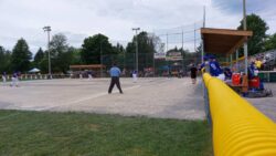 A fastpitch softball match. Team Quebec is in the outfield in blue jerseys. The Delisle Pride are wearing black jerseys and are batting. Spectators sit in the stands.