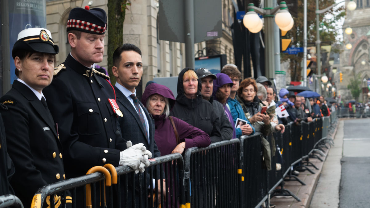 People standing behind metal barricades look towards the parade with gloomy expressions.