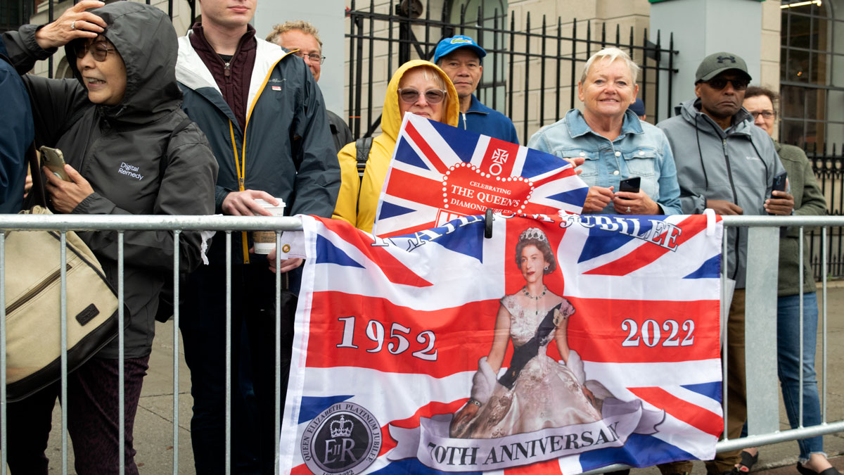 A woman in a yellow coat stands behind a flag with an image of a younger Queen Elizabeth emblazoned on it.