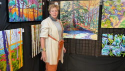 Female artist stands in front of colourful paintings of trees.