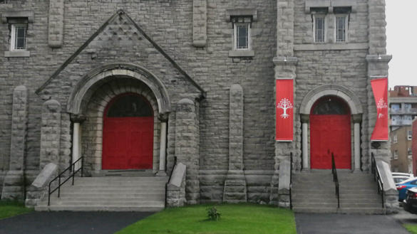 The front facade of St. Brigid's former church after The United People of Canada began using the building as an "Embassy." The doors have been painted red, and red flags can be seen on the door to the right.