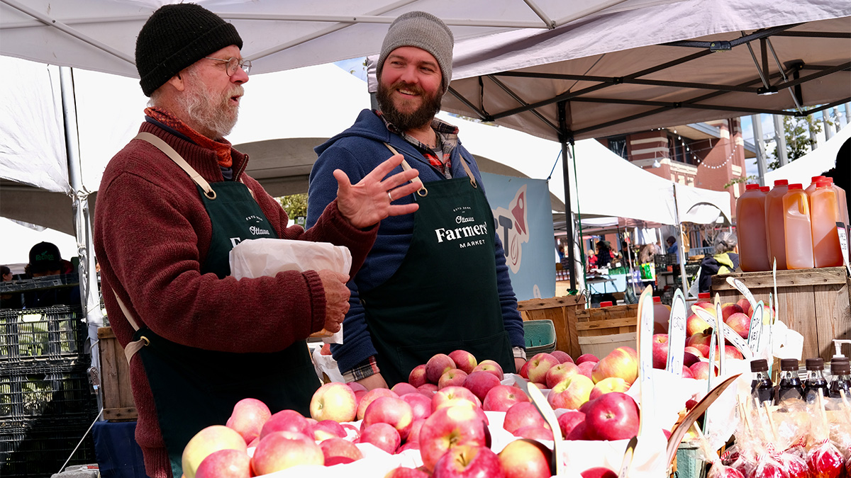 Man looks at older man who is speaking at a farmer's market booth covered in apples.