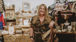 A blonde woman leans on a shelf of vintage pottery and decor items in front of the Vintage Booth she runs. In the background is clothing racks and vintage home wares