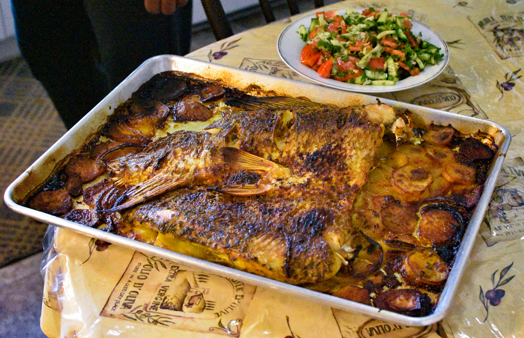 A fish has been cut in half, laid on a tray and cooked in turmeric with potatoes, onions and oil. Beside it is a cucumber and tomato salad with pomegranate molasses.