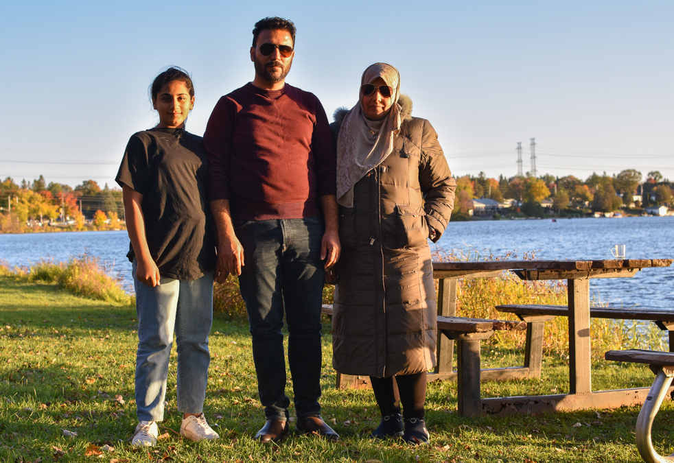 Omar Masri, his wife and daughter stand in Mooney's Bay beside the Rideau River and a couple picnic tables