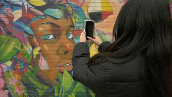 Girl stands in front of mural with phone in her hand to take picture of it.