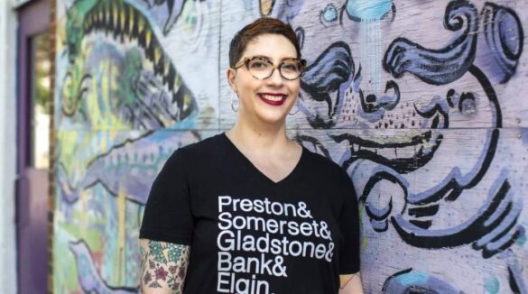 Ariel Troster wears a shirt that says "Preston & Somerset & Gladstone & Bank & Elgin" while standing in front of a graffitied wall.