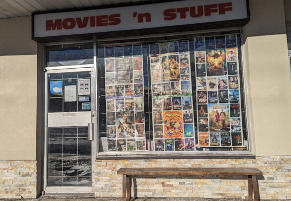 The midday sun casts a shadow on Movies 'n Stuff's sign and contrasts the sun's warmth on the stone wall. On even closer inspection, snow reflects in the store's glass door, and dozens of colourful movie posters fill the windows. The open sign flashes welcomely to customers.