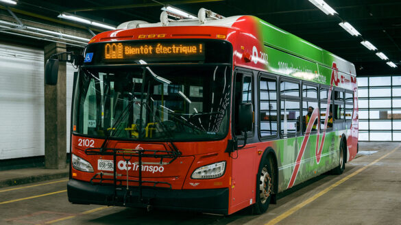 Inside an Ottawa garage is one of OC Transpo’s new electric buses. Its red, white, and green exterior shine under overhead lighting. The front of the bus has a digital banner that reads “Bientôt électrique!”
