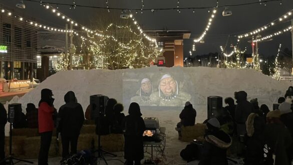 A crowd of moviegoers all dressed in their winter coasts gathered in Landsdowne park watching the short film Angakusajaujuq: The Shaman's Apprentice being projected onto the handcrafted snow screen at night, so it is dark outside.