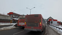 A red and white diesel-powered OC Transpo bus idles near the sidewalk on a cold and cloudy winter afternoon. No one is on the bus, nor does anyone get on it, yet it remains pumping exhaust into the air. Unperturbed by the familiar sight, another bus drives past it.