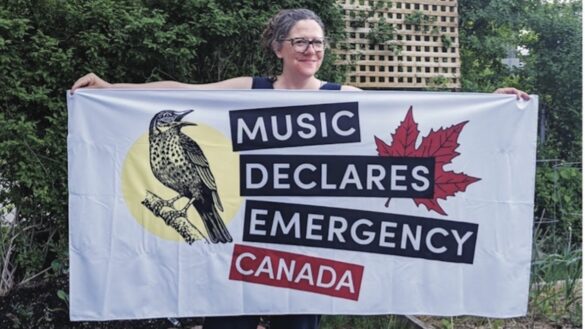 Kimberly Fry proudly poses outside with a flag, reading "Music Declares Emergency Canada"