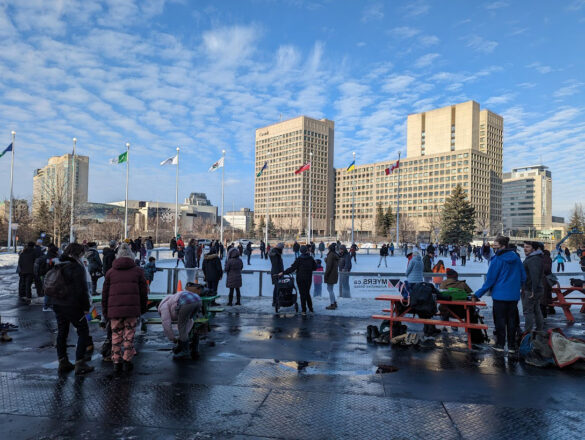 Bundled up in winter gear, Ottawa’s 2SLGBT+ residents mingle and skate on the Rink of Dreams in front of City Hall. The sun creates puddles on the ground and brightens buildings downtown while flags blow in the wind. Those not actively moving about on the rink sit on benches and watch on.