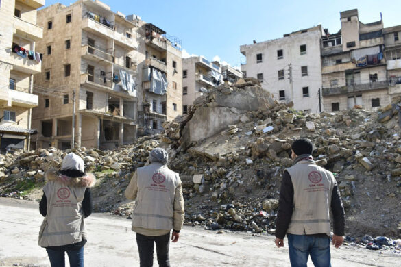 Three people stand facing the rubble from the earthquakes. Buildings in the distance.