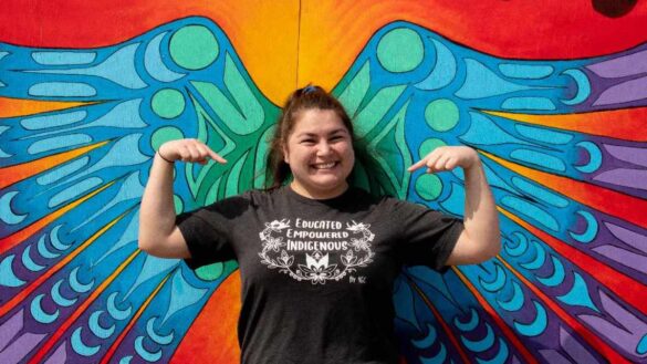 Paula MacDonald flexing her arms in a strong pose in front of blue and purple ombre Indigenous style wings painted on a wall.