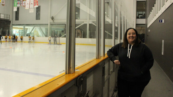 A woman leans up against the boards of a hockey rink, smiling at the camera.