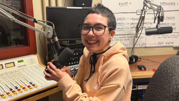 Gonzalez, a student at uOttawa, sits in a radio studio at CHUO FM. They are smiling at the camera.