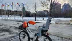 Bike stands in front of City Hall flags in the winter. It is equipped with a children's seat and gloves attached to the handles.