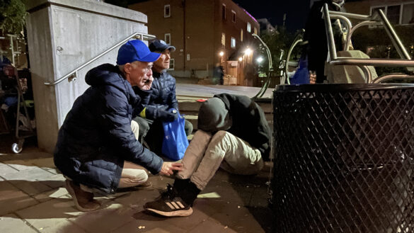 A man wearing a blue cap crouching down beside a man with his head in his knees who is suspected of an overdose. The man in the blue cap is on the phone to 911 and has his hand placed on the other man's knee.