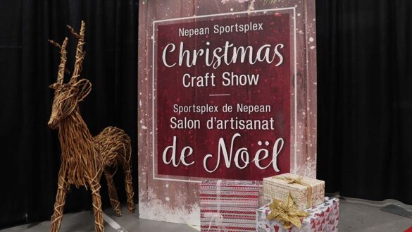 A red and white sign with the words "Nepean Sportplex Christmas Craft Show" written in the middle in French and English. A wooden reindeer is on the left of the sign. There are red and white presents are stacked in the bottom right corner of the sign.