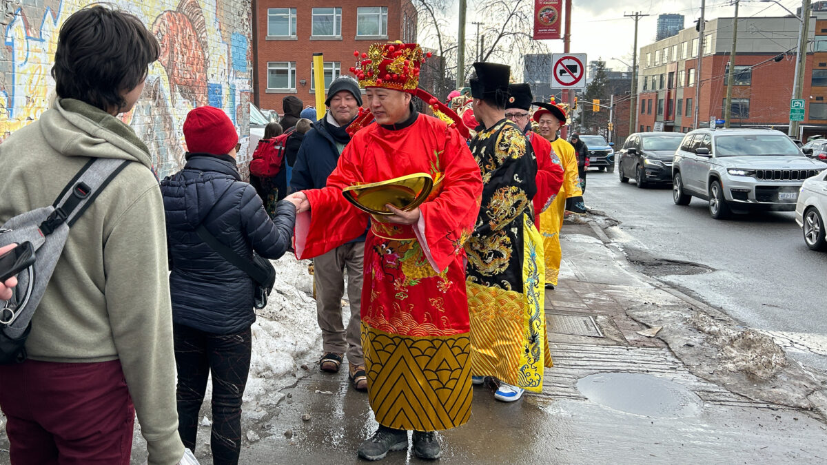A man wearing a red outfit and hat hands out red envelopes to people on the sidewalk.