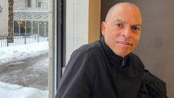 Photo of Robin Browne sitting in a seat by a window in Planet Coffee. Browne is captured wearing a black sweater in the right section of the photo and to the left is a view of the window and outside of the restaurant.