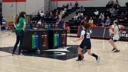 Two girls in basketball uniforms dribble up the court while holding pieces of recyclable waste.