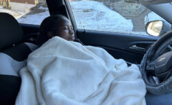 Woman covered in blanket sleeps in driver's seat of car, snow visible outside